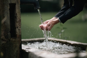 Zwei Hände unter fließendem Wasser, welches aus einem alten, hölzernen Brunnen fließt. Die Umgebung ist ein ruhiger, grüner Garten.