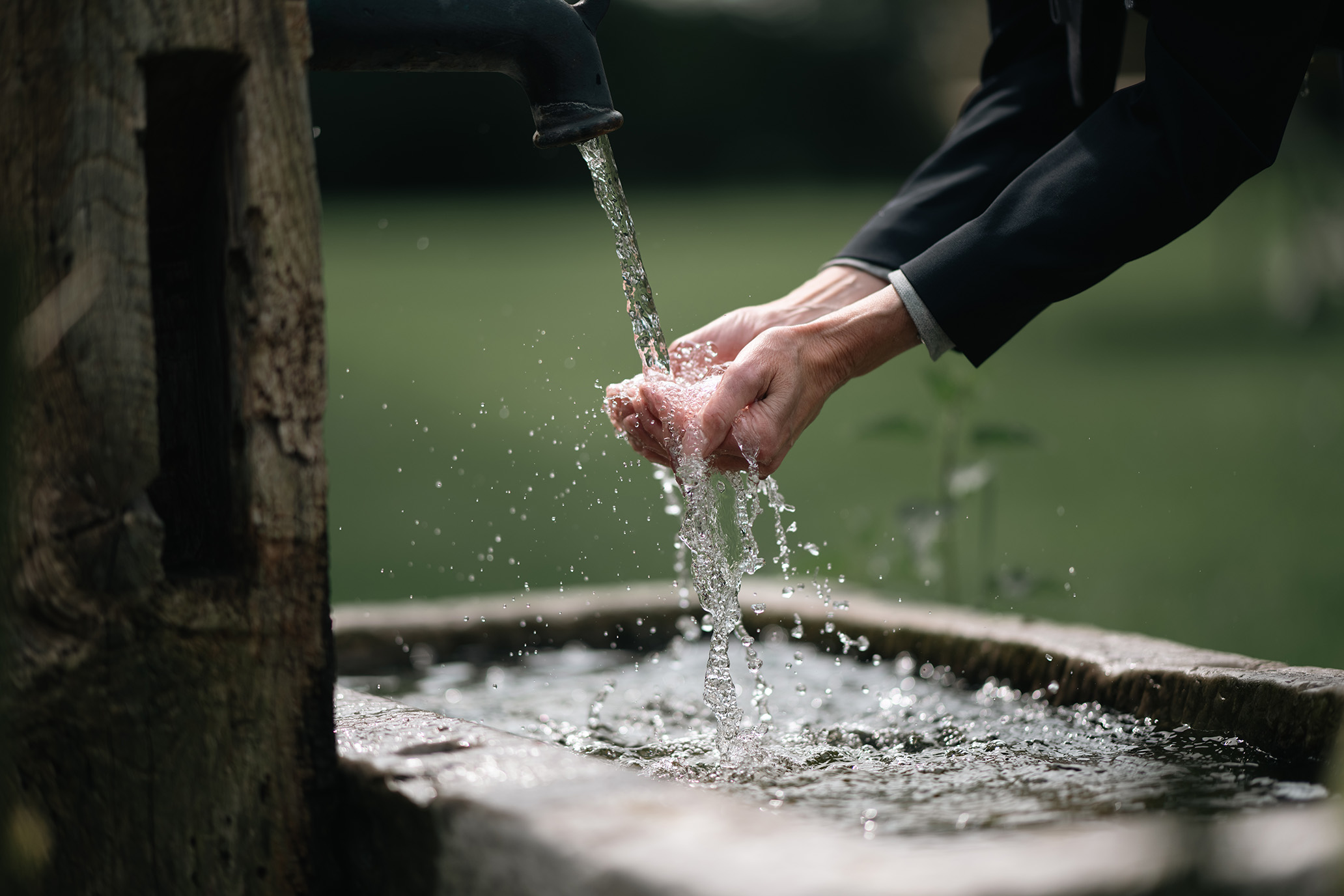 Zwei Hände unter fließendem Wasser, welches aus einem alten, hölzernen Brunnen fließt. Die Umgebung ist ein ruhiger, grüner Garten.